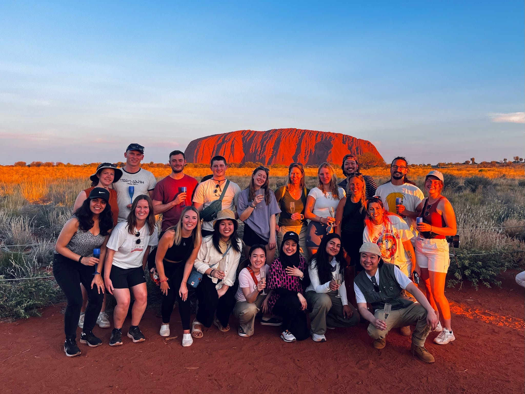 A group of tourists posing together with Uluru in the background during sunset.