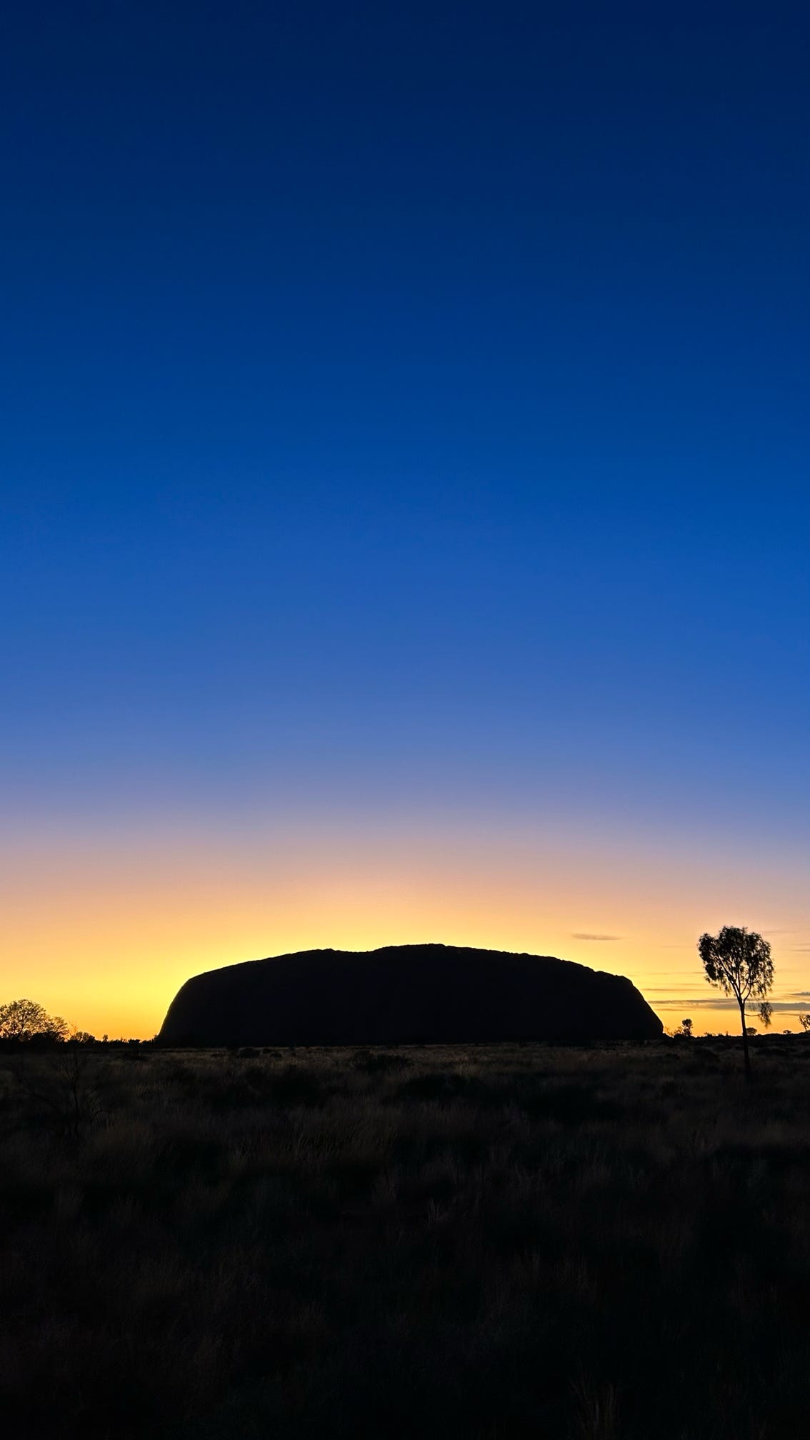 The silhouette of Uluru stands majestically against the twilight sky, transitioning from a golden hue to deep blue, capturing the serene beauty of the Australian outback.