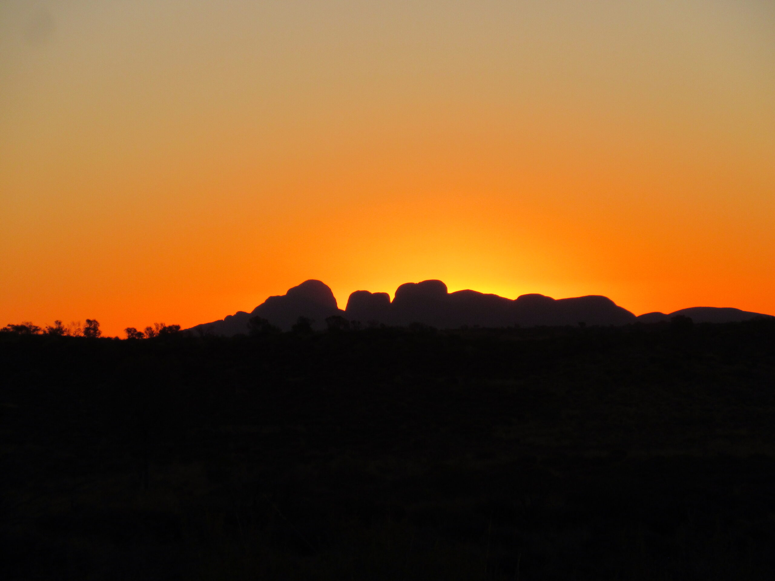 A stunning sunset behind Kata Tjuta (The Olgas), creating a dramatic silhouette against a vibrant orange sky.