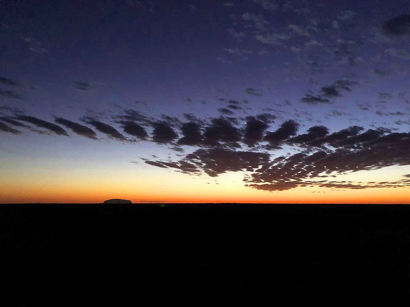 Uluru stands majestically silhouetted against a stunning sunset, with dramatic clouds stretching across the sky. This serene moment captures the natural beauty and tranquility of the Outback at dusk.