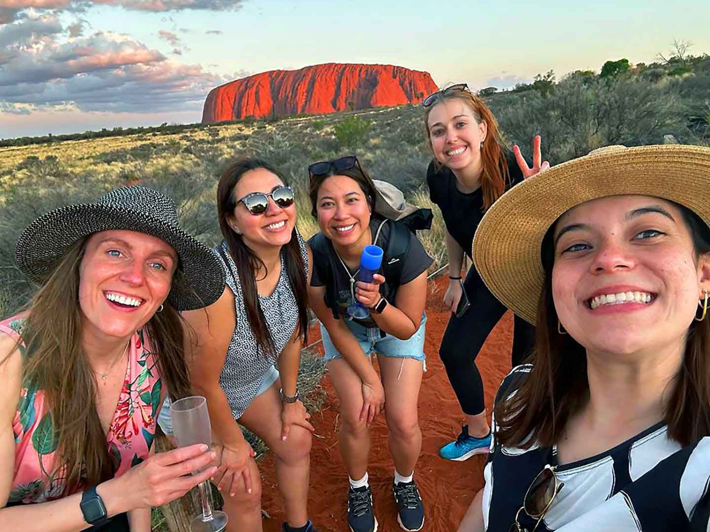 A joyful group of Mulgas Adventures tourists posing for a selfie with Uluru's majestic presence in the background, bathed in the warm hues of the setting sun.