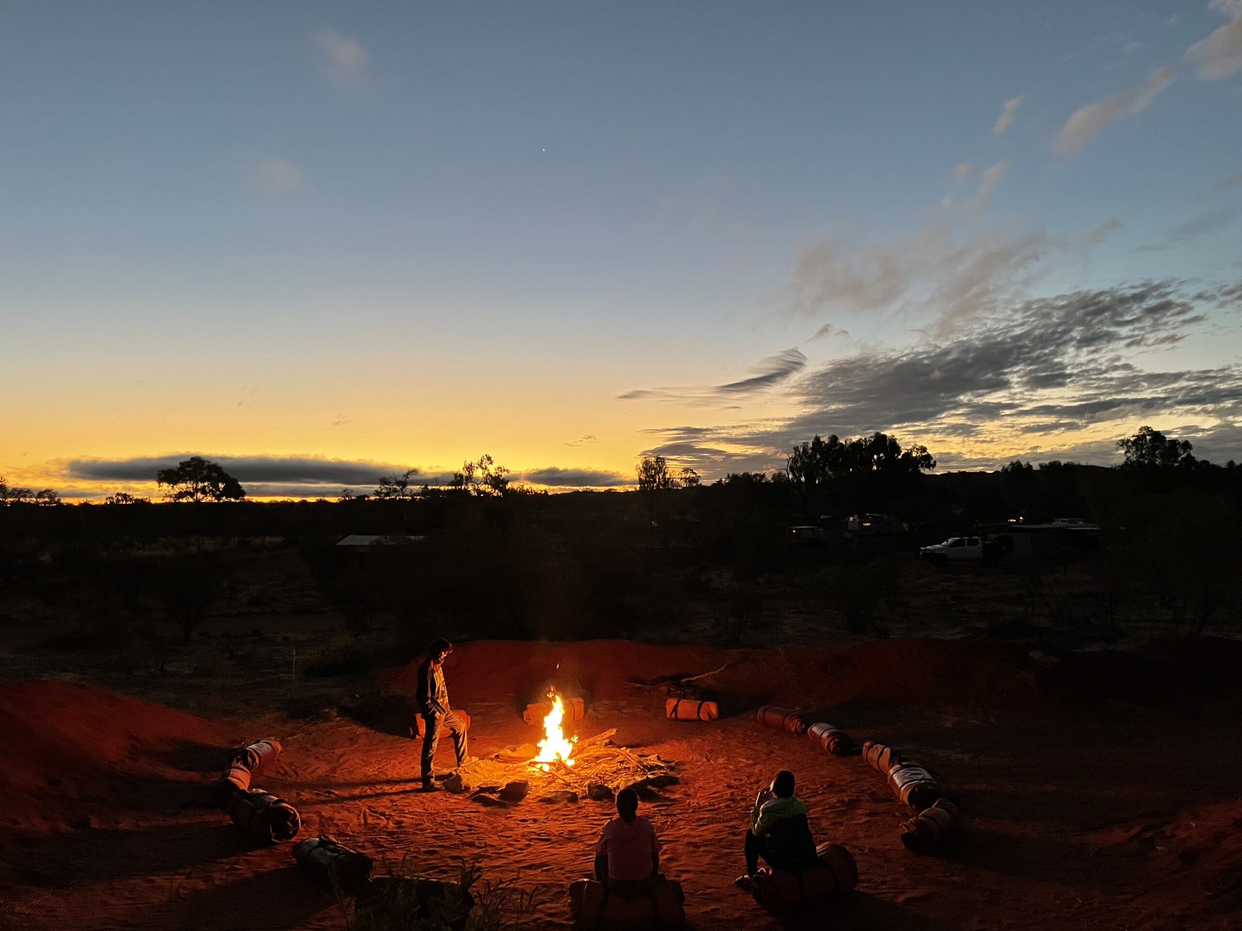 A serene moment at dusk as tour members gather around a campfire, with swags laid out in a circle and the sky painted with the colors of the setting sun.