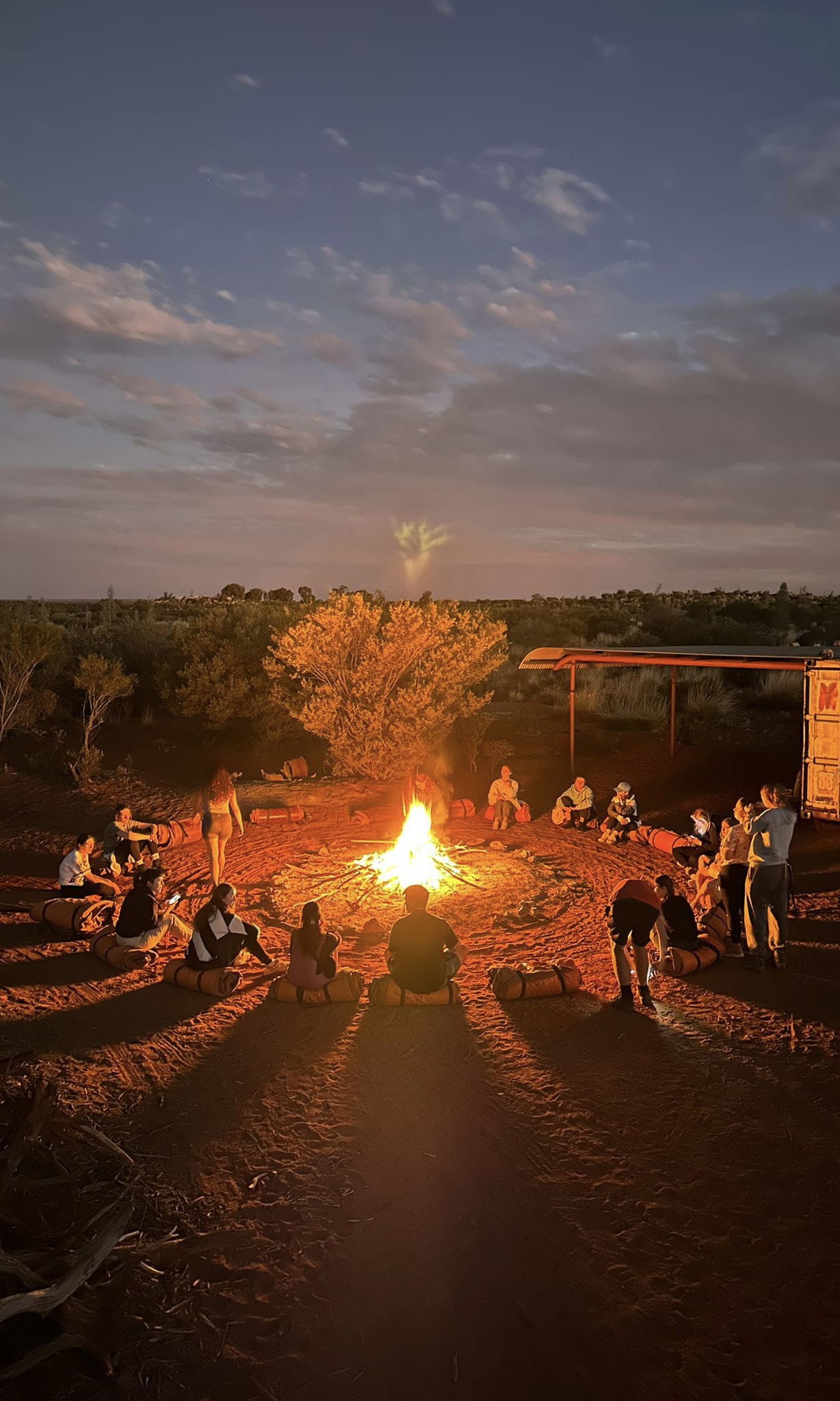 A tour group from Mulgas Adventures gathers around a campfire at dusk, enjoying the serene atmosphere and camaraderie under the twilight sky.