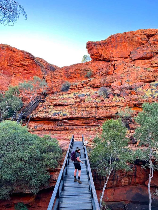 Mulgas Adventures tour guide Laura leads a hike across a bridge at Kings Canyon, showcasing the dramatic red cliffs and lush greenery of this iconic Australian landscape.