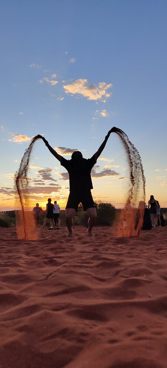 A Mulgas Adventures tour group member joyfully tosses sand into the air at sunset, creating a striking silhouette against the colorful sky.