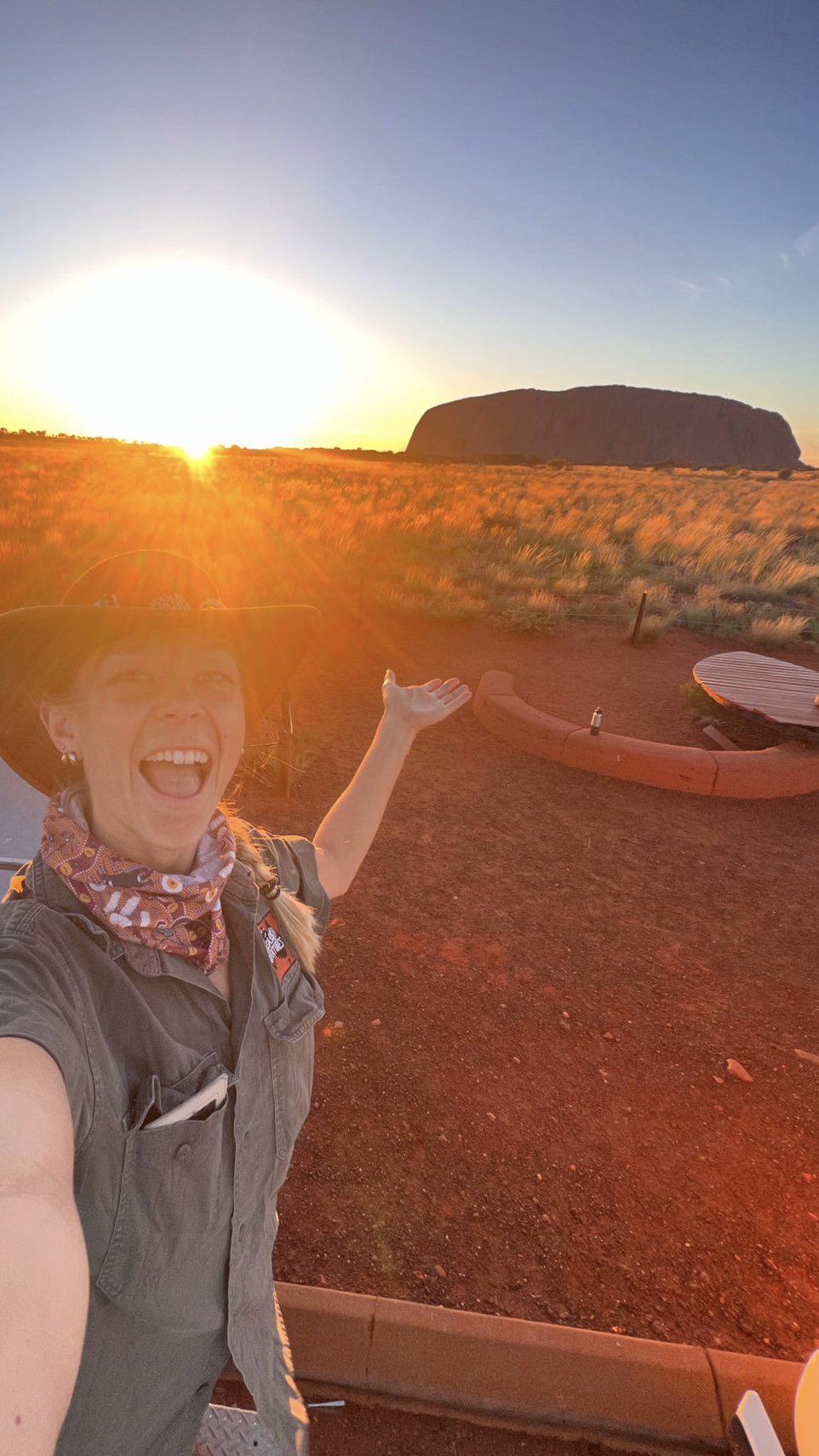 A cheerful tour guide welcomes travelers to experience the stunning sunset at Uluru, with the sun setting behind the iconic rock formation, creating a golden glow over the landscape.