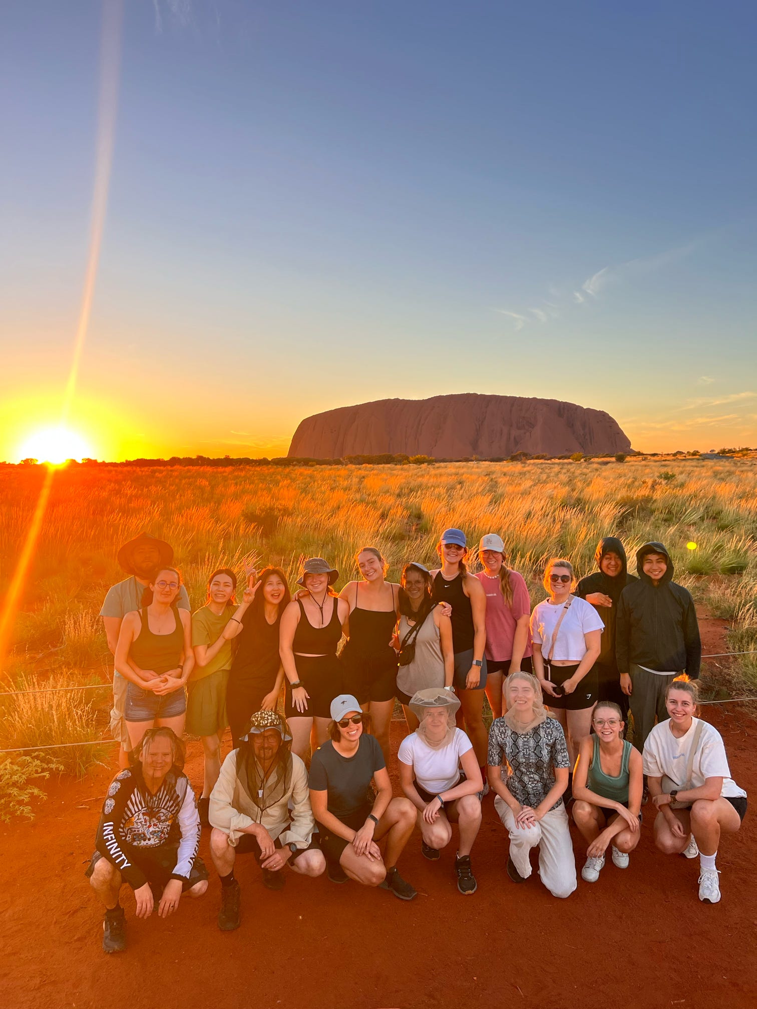 A group of excited travelers posing in front of Uluru during a stunning sunrise, capturing the iconic beauty of the Australian outback with Mulgas Adventures.