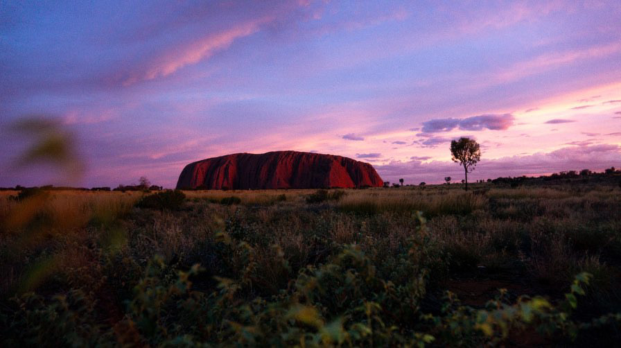 The majestic Uluru is bathed in the warm hues of sunset, set against a vibrant sky in Central Australia, capturing the natural beauty of this iconic landmark. Photo credit: Ben Lange. Instagram: @benlangeee