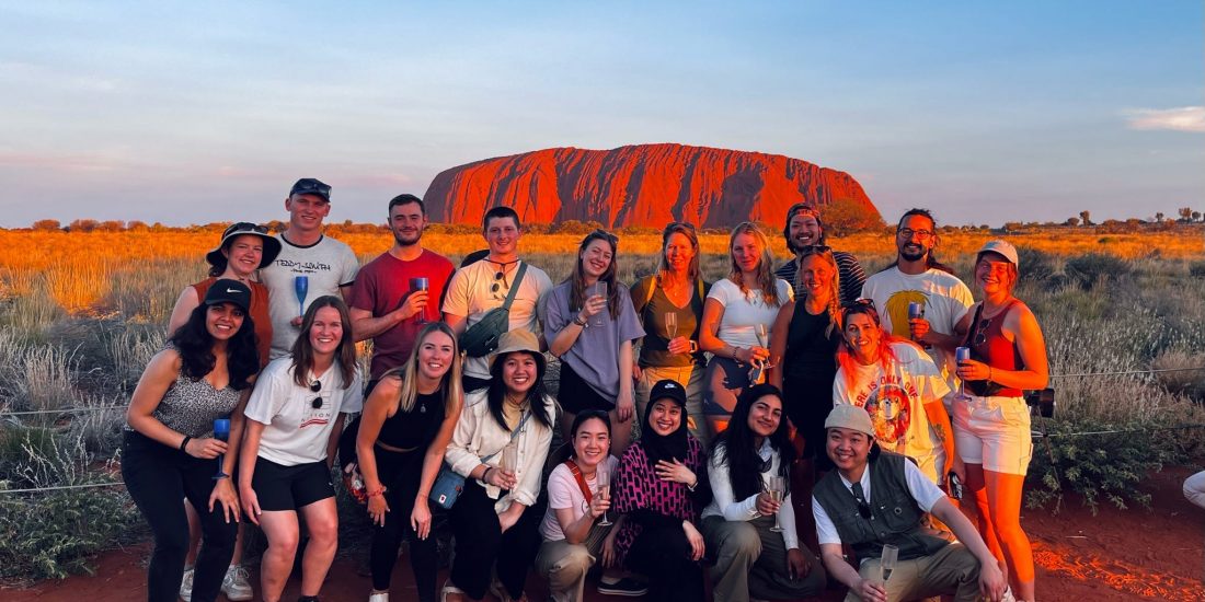 A group of tourists posing together with Uluru in the background during sunset.