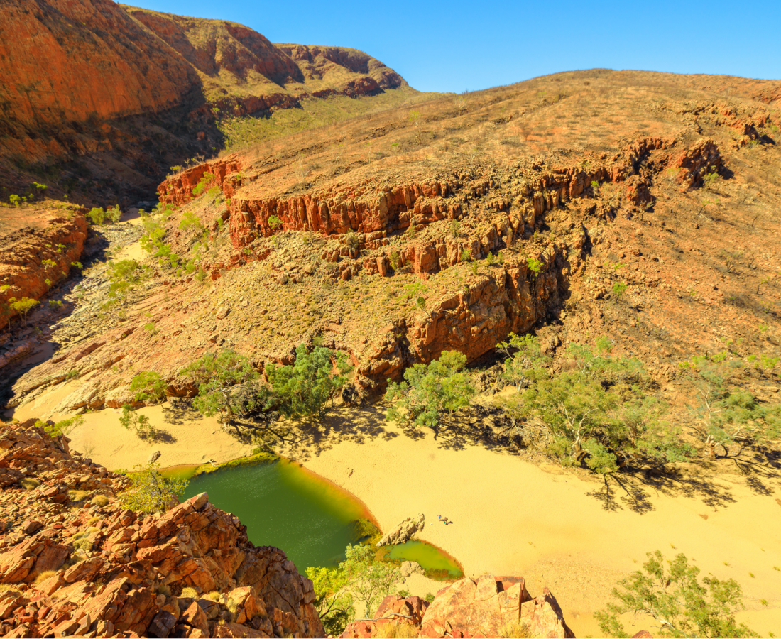 Aerial view of Ormiston Gorge with a sandy beach and waterhole surrounded by rugged red rock cliffs.