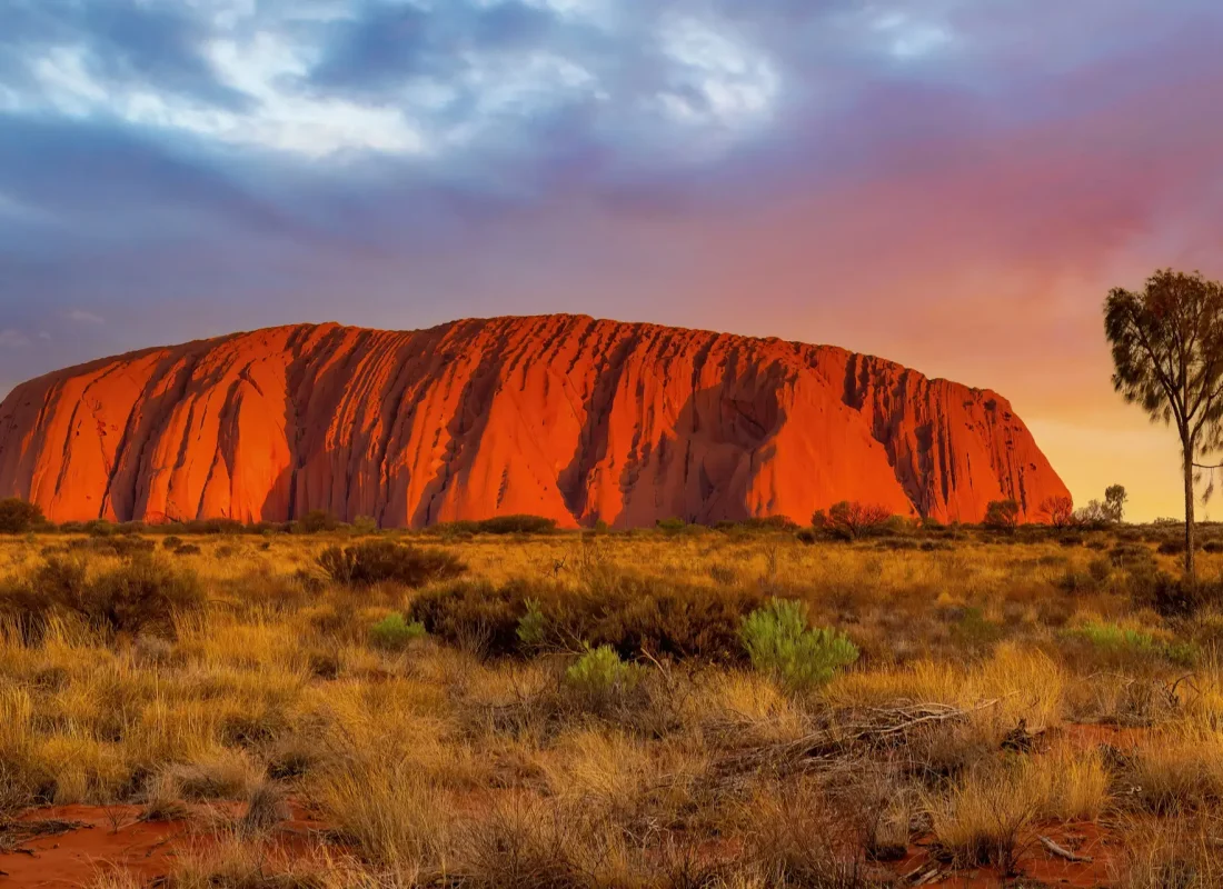 Uluru at sunset, glowing red against a backdrop of a colorful sky with scattered clouds.